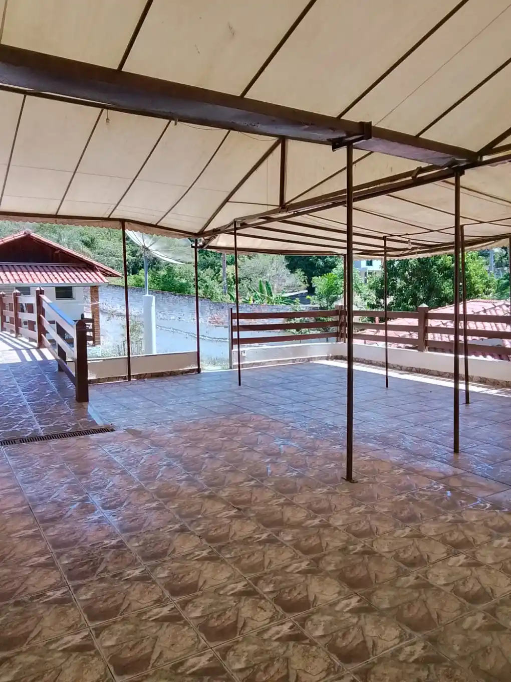 Área coberta com piso cerâmico e cobertura de lona, aberta para a vegetação.